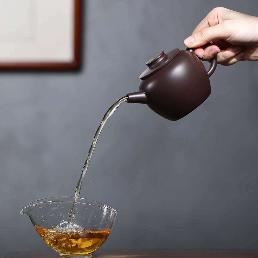 Handmade Yixing purple sand teapot pouring tea into a glass bowl, showcasing fine craftsmanship and traditional tea preparation.