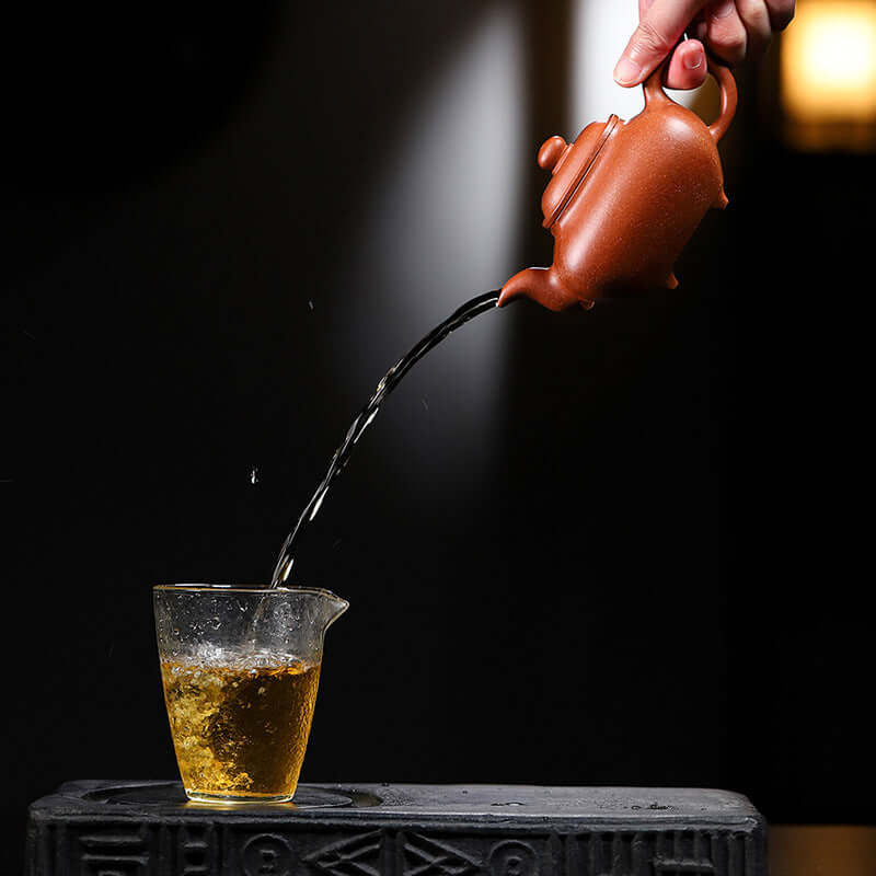 Handmade Yixing purple sand teapot pouring tea into a glass cup, showcasing artisan craftsmanship.