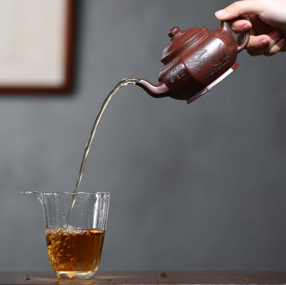 Handmade Yixing purple sand teapot pouring tea into glass cup, showcasing craftsmanship and elegance.