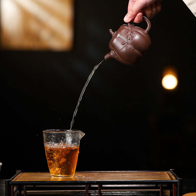 Handmade Yixing purple sand teapot pouring tea into a glass cup, showcasing traditional craftsmanship and elegance.
