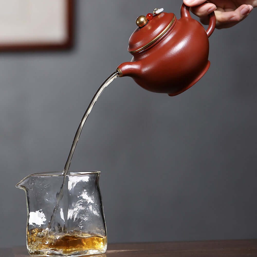 Handmade Yixing purple sand teapot pouring tea into a glass cup, showcasing its unique red clay design and craftsmanship.