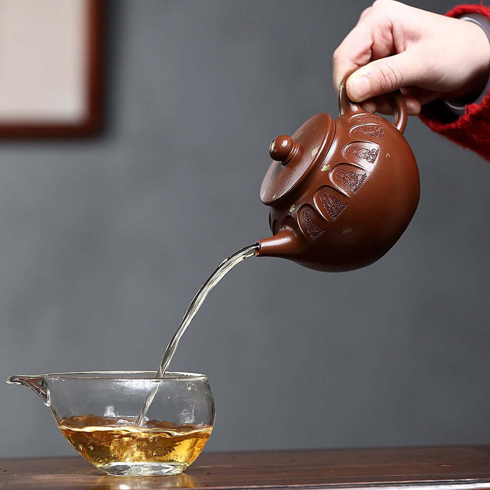 Handmade Yixing purple sand teapot pouring tea into a glass cup, showcasing Dunhuang heritage craftsmanship.