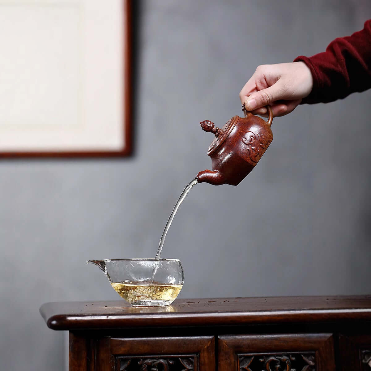 Handmade Yixing purple sand teapot pouring tea into a glass cup, showcasing authentic craftsmanship.