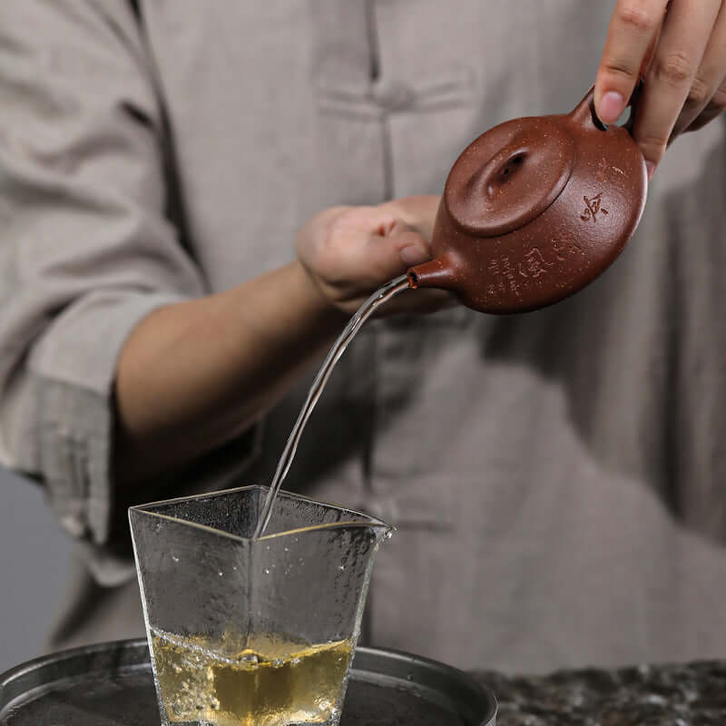 Handmade Yixing purple sand teapot pouring tea into a glass cup, showcasing craftsmanship and elegance.