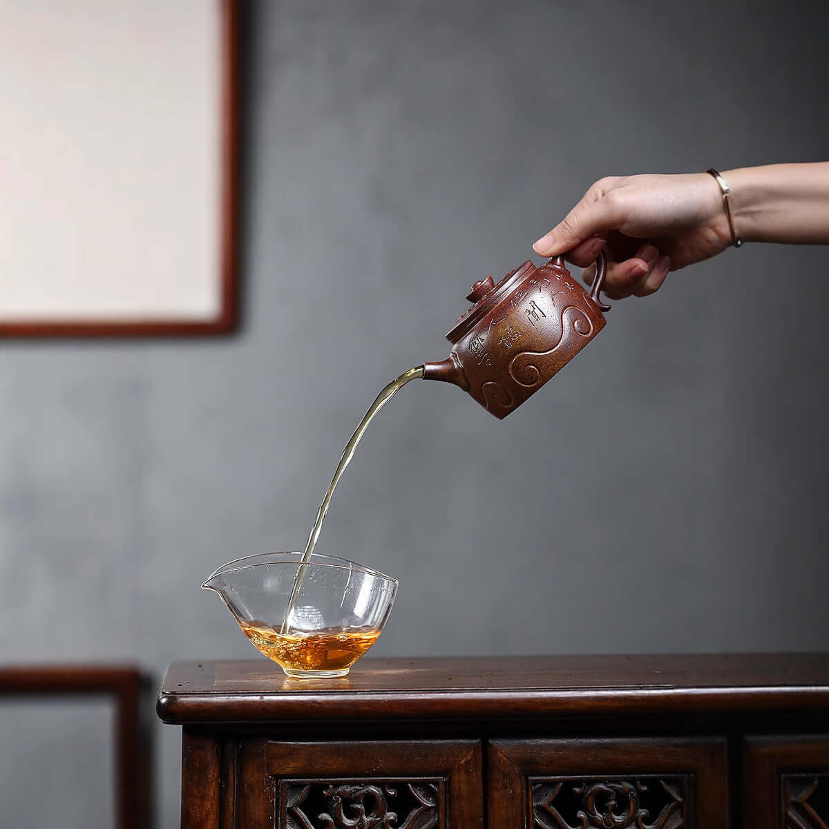 Handmade Yixing purple sand teapot pouring tea into a glass cup on a wooden table, showcasing traditional craftsmanship.