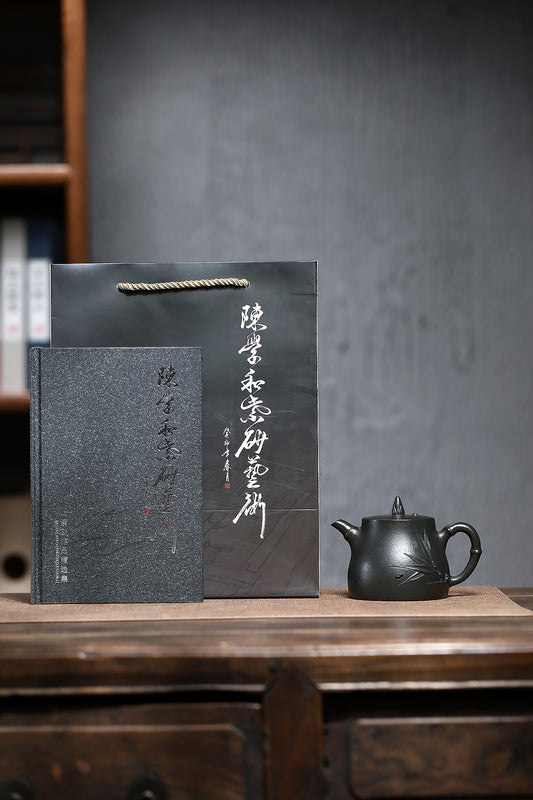 Handmade Yixing purple sand teapot next to an art book and elegant gift bag on rustic wooden table.