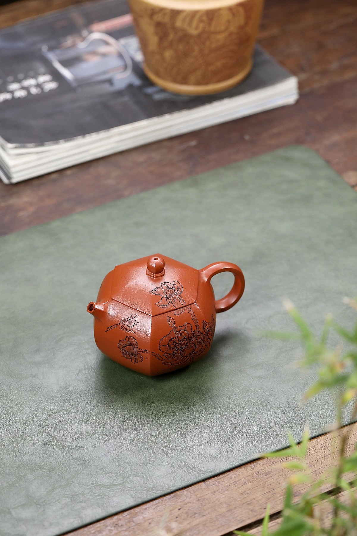 Handmade Yixing purple sand teapot, featuring delicate lotus engraving, on a green mat with books in the background.
