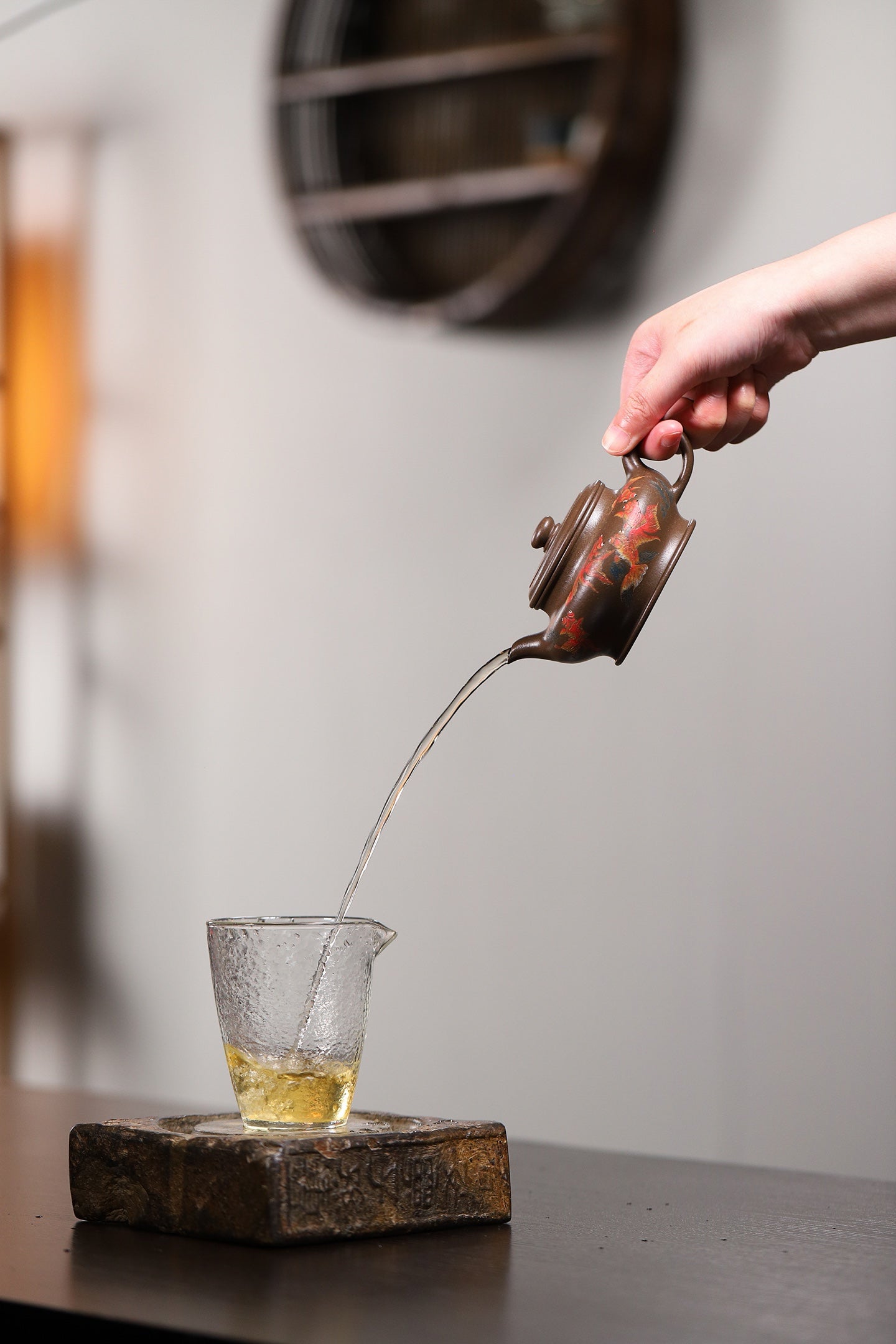 Hand pouring tea from a handmade Yixing purple sand teapot into a glass cup, showcasing elegant craftsmanship.