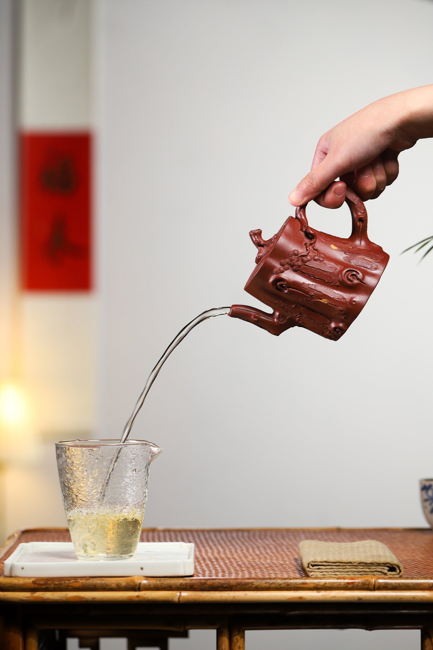 Hand pouring tea from a handcrafted Yixing purple sand teapot into a glass cup on a bamboo tray.