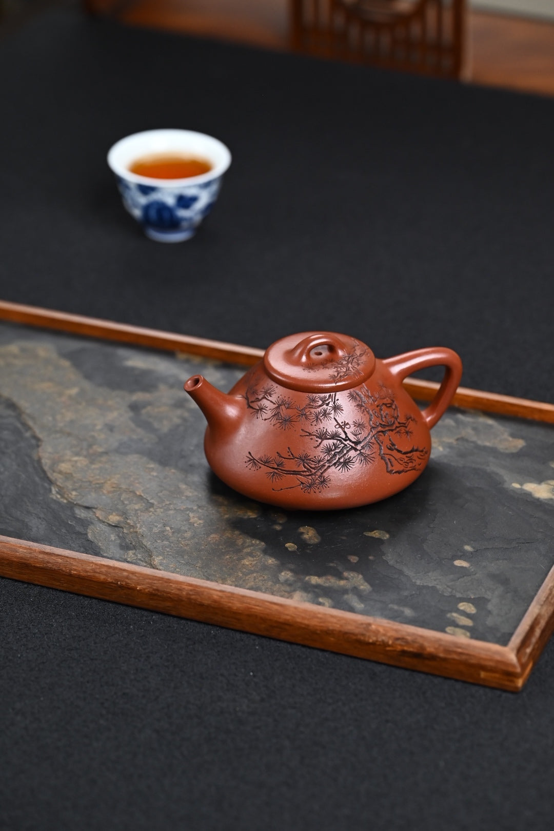 Handmade Yixing purple sand teapot on a stone tray with a cup of tea in the background, showcasing traditional craftsmanship.