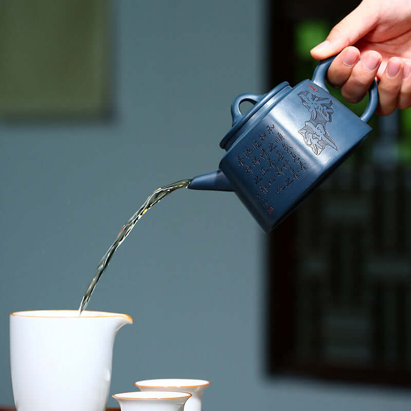 Hand pouring tea from a handmade Yixing purple sand teapot into a white cup. Artisanal craftsmanship evident in design.