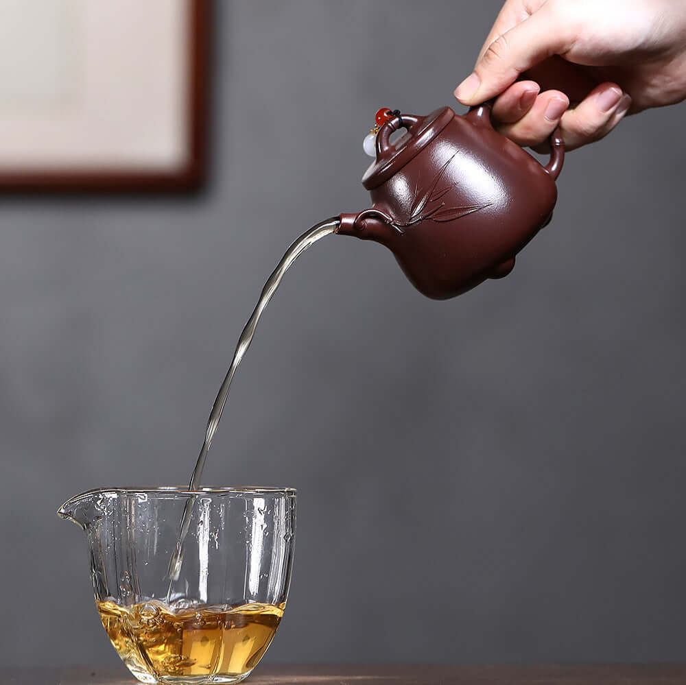 Handmade Yixing purple sand teapot pouring tea into glass cup, showcasing traditional craftsmanship and elegance.