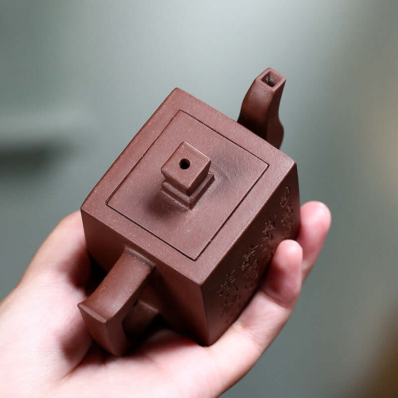 Handmade Yixing purple sand teapot held in hand, showcasing its square design and intricate details.