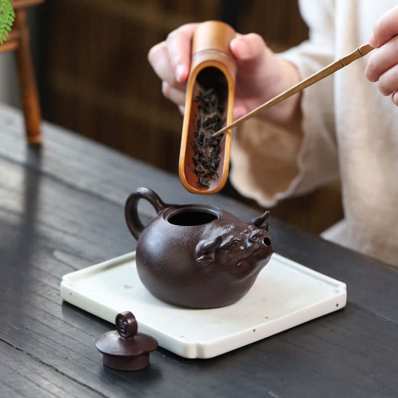 Handmade Yixing purple sand teapot shaped like a bull, being filled with loose tea leaves on a wooden table.