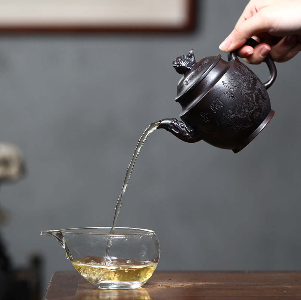 Handmade Yixing purple sand teapot pouring tea into a glass cup, showcasing traditional craftsmanship and artistry.