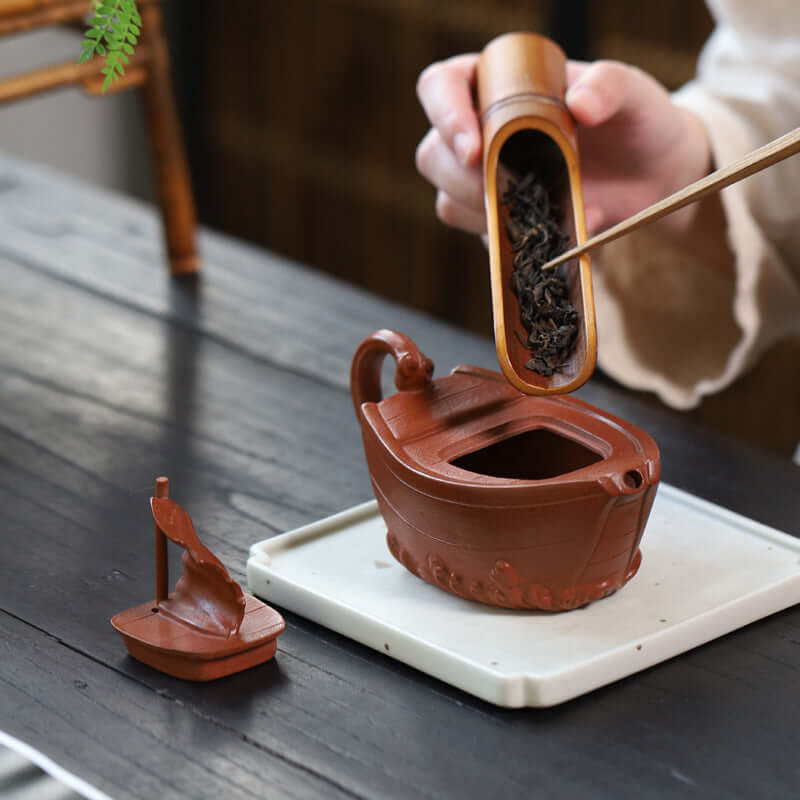 Handmade Yixing purple sand teapot with unique boat design, filled with tea leaves, on a wooden table.