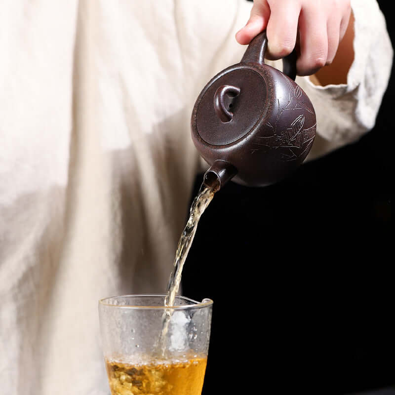 Handmade Yixing purple sand teapot pouring tea into a glass cup, showcasing elegant craftsmanship and traditional tea culture.