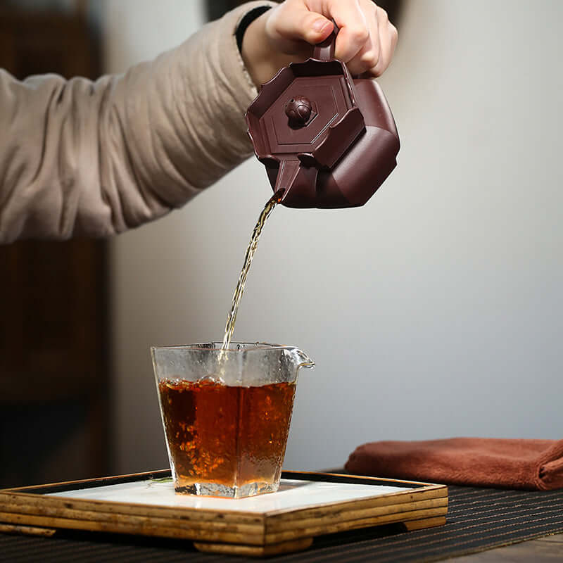 Hand pouring tea from a handmade Yixing purple sand teapot into a glass cup, showcasing artisanal craftsmanship.
