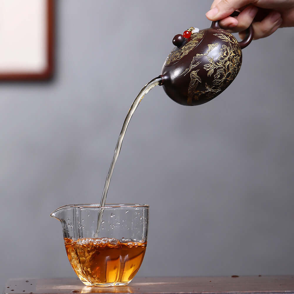 Handmade Yixing purple sand teapot pouring tea into a glass cup, showcasing intricate floral design and craftsmanship.