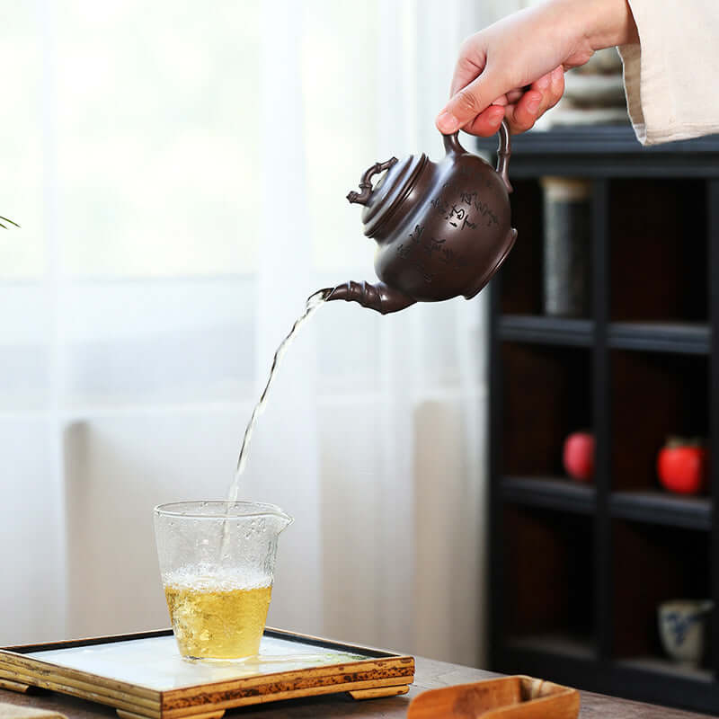 Handmade Yixing purple sand teapot pouring tea into a glass cup, showcasing elegant craftsmanship and functionality.