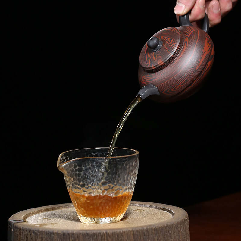 Handmade Yixing purple sand teapot pouring tea into a textured glass cup on a wooden surface.