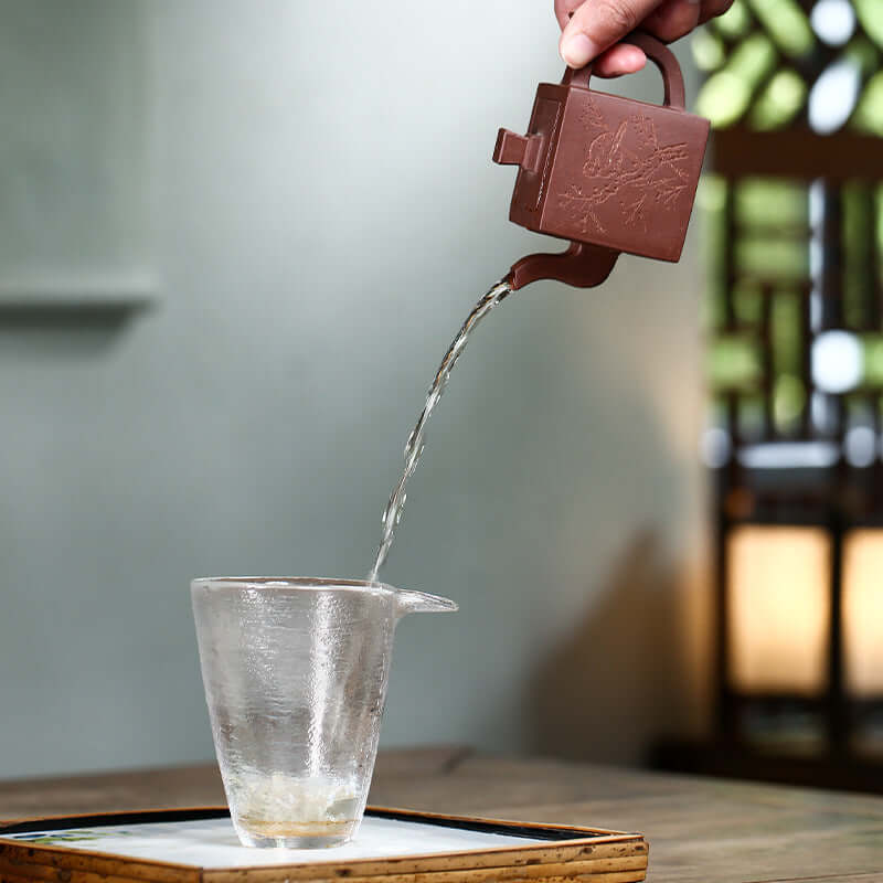 Handmade Yixing purple sand teapot pouring tea into a glass cup, showcasing craftsmanship and traditional tea ceremony.