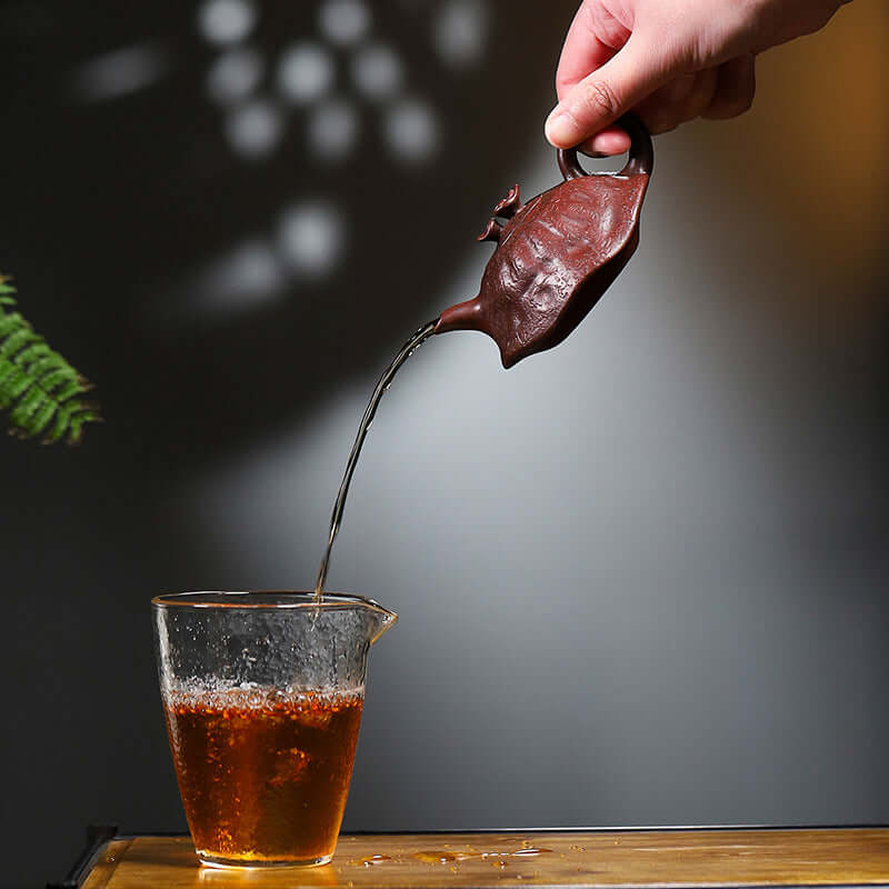 Handmade Yixing purple sand teapot pouring tea into a clear cup, showcasing fine craftsmanship and rich color.