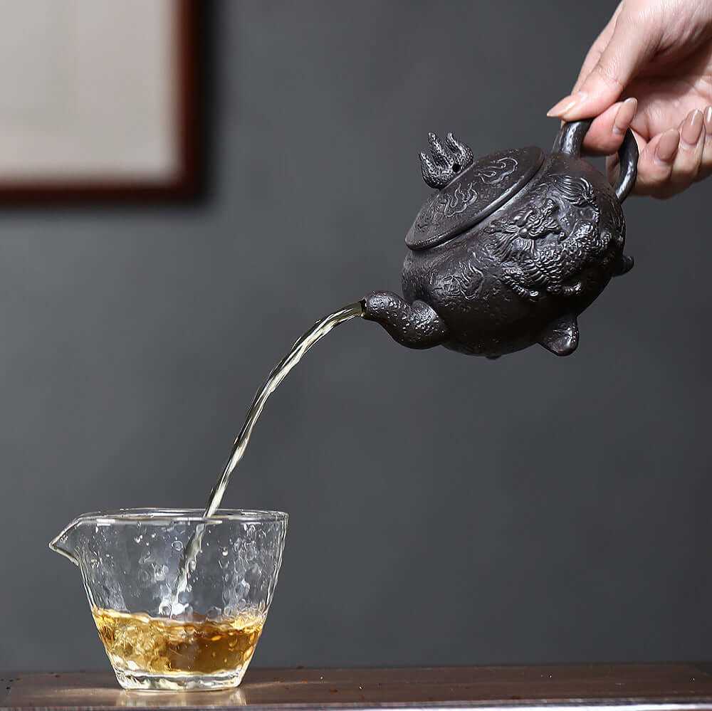 Handmade Yixing purple sand teapot pouring tea into a glass cup, showcasing intricate craftsmanship and design.