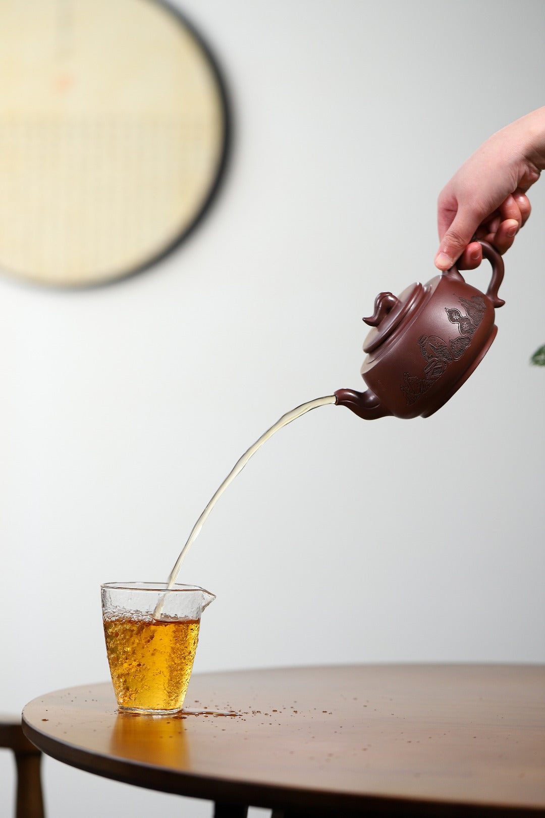 Handmade Yixing purple sand teapot pouring tea into a glass cup on a wooden table.