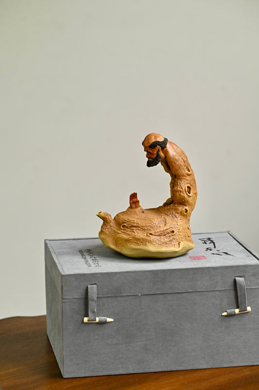 Handmade Yixing purple sand teapot depicting a meditative figure, showcased on a gray gift box.