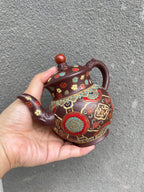 Yixing purple clay teapot with enamel decoration held by hand against a textured gray background.